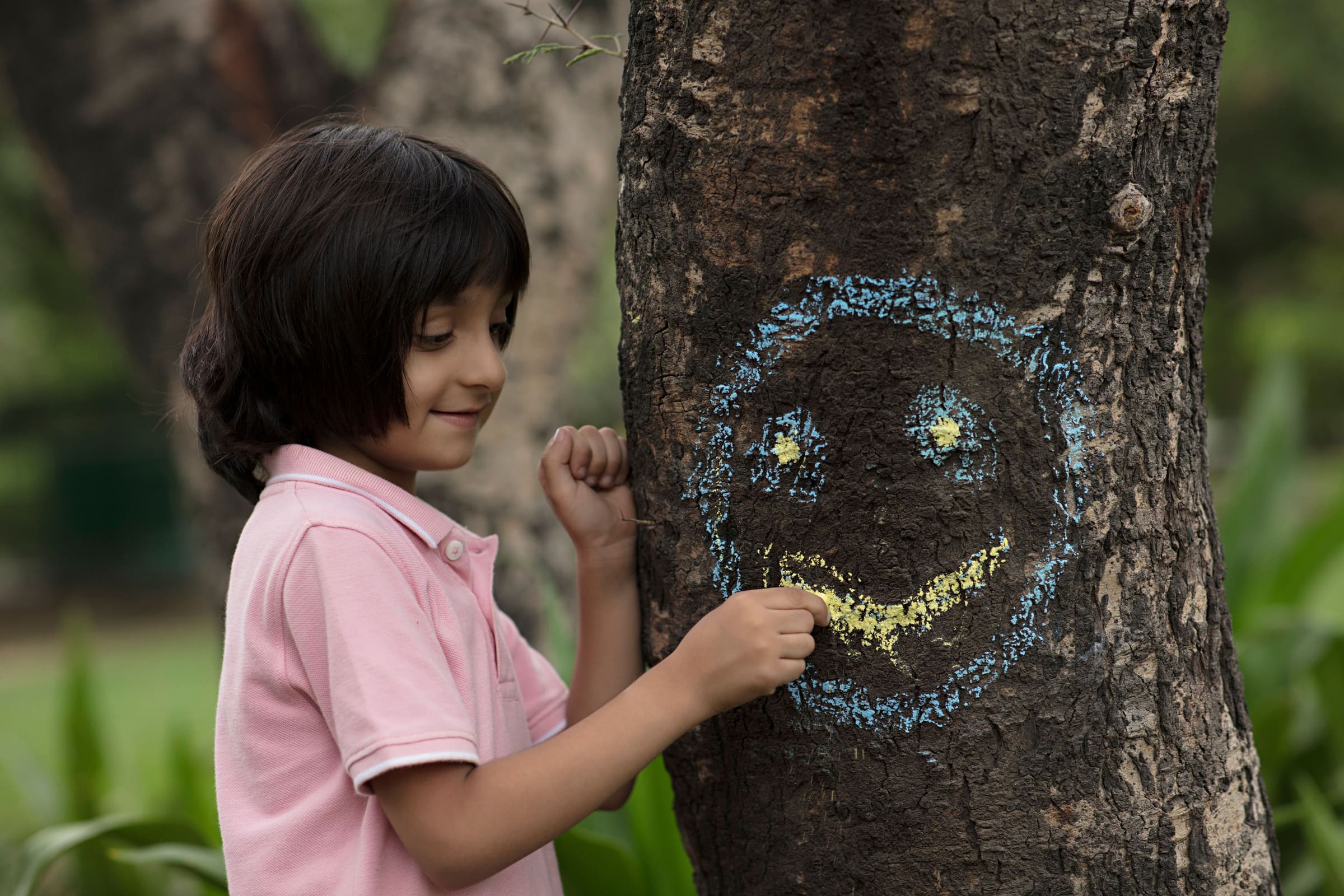 A smiling child chalking a bright smiley face onto a tree trunk in a green park