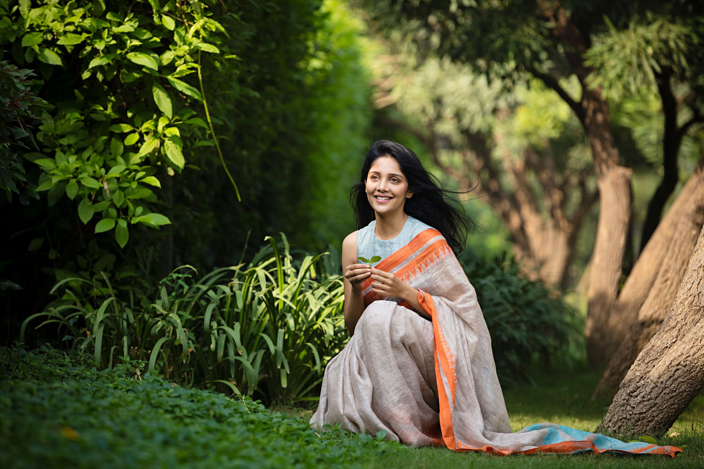 A woman in a saree sitting in a sunlit garden, smiling as she cups a young seedling in her hands